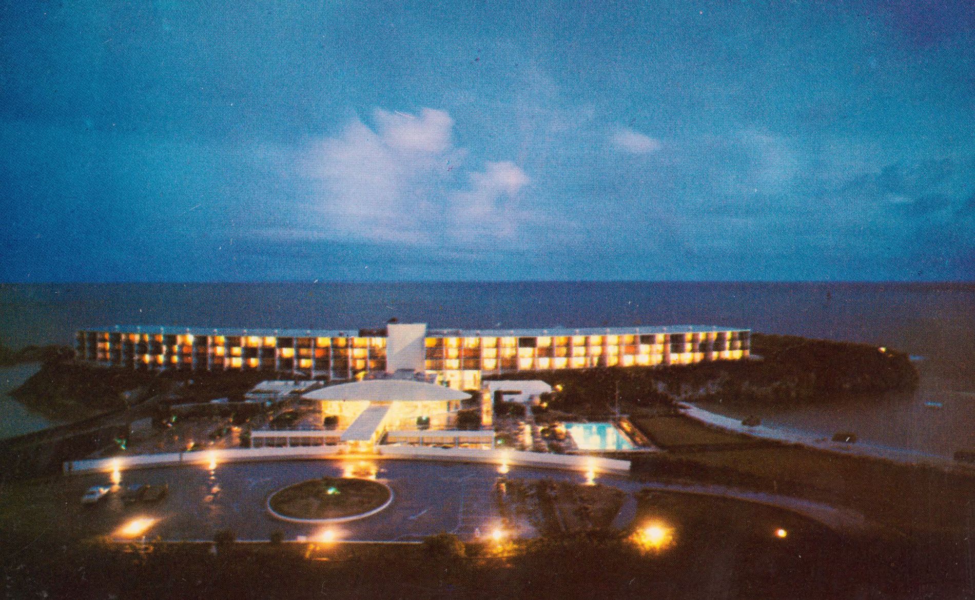 A vintage postcard of a seaside hotel and its pool lit up at night, under a blue evening sky